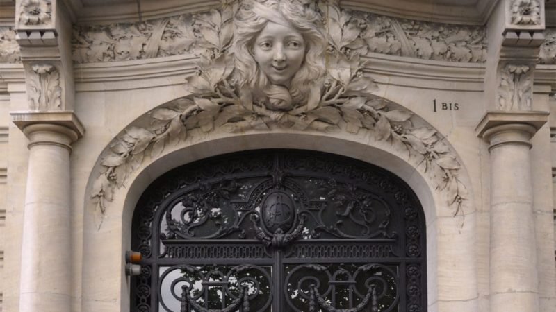 Un portail en fer forgé orné est placé dans une arche en pierre décorée de sculptures complexes. Au-dessus de la porte, un relief en pierre présente une sculpture détaillée d'un visage de femme entouré de feuilles. Le bâtiment, marqué du numéro « 1 BIS », dégage un air de grandeur historique qui fait écho à la solidité des garanties prêt immobilier.