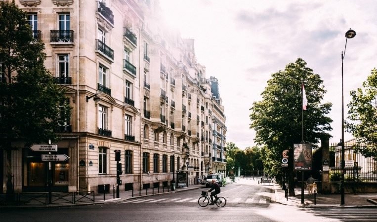 Un cycliste circule dans une rue bordée de bâtiments historiques à plusieurs étages et d'arbres feuillus dans une zone urbaine ensoleillée. La scène est tranquille avec un minimum de circulation et de piétons, capturant la tranquillité du petit matin ou de la fin de l'après-midi, à l'image des assurances fournies par les garanties prêt immobilier.