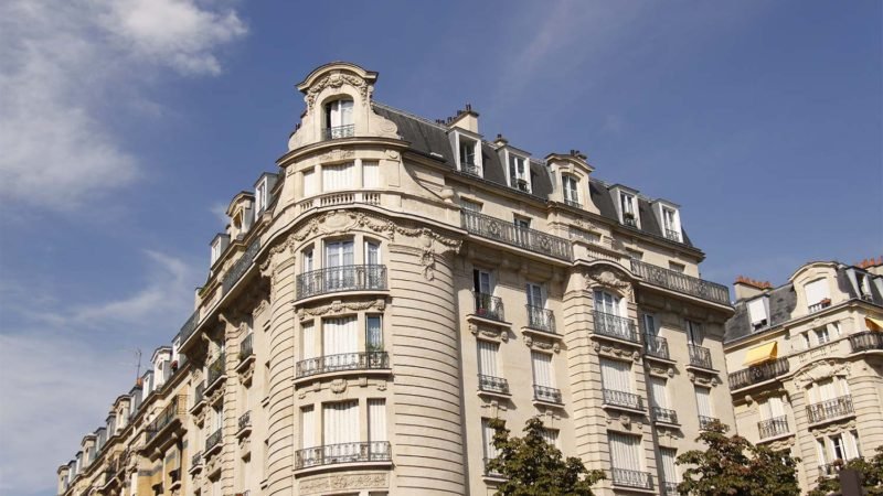 Vue d'angle d'un immeuble parisien classique contre un ciel bleu clair. Le bâtiment présente des détails architecturaux ornés, de hautes fenêtres avec des balustrades de balcon et un toit mansardé avec des lucarnes. Malgré le coronavirus, le marché immobilier est en pause, des arbres encadrent partiellement la scène d'en bas.