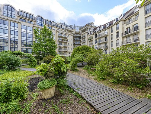 Vue sur un jardin intérieur dans un complexe résidentiel comprenant plusieurs immeubles d'appartements modernes. Le jardin comprend des plantes vertes luxuriantes, des arbustes, une variété d'arbres et une passerelle en bois. De hautes fenêtres et balcons sont visibles sur les bâtiments environnants.