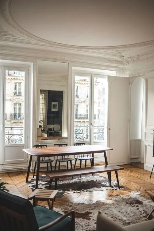 Une pièce élégante et ensoleillée avec parquet comprend une longue table à manger avec des bancs, entourée de murs lambrissés blancs et de grandes fenêtres cintrées laissant entrer la lumière naturelle. Un miroir reflète l'intérieur et un tapis en peau de vache ajoute une touche de charme rustique : une trouvaille parfaite pour tout chasseur d'appartement à Paris.