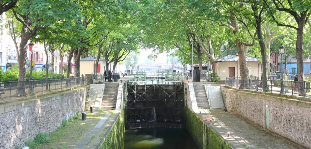 Une écluse sereine entourée d'arbres verts luxuriants dans un parc urbain définit le décor, rappelant le Chasseur d'appart Paris 10. Des marches en pierre mènent au canal, où l'eau calme passe à travers les portes de l'écluse. Les piétons des deux côtés de l’écluse profitent de l’environnement paisible.