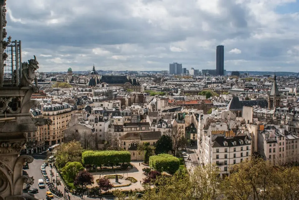 Vue de Paris depuis un point de vue surélevé, montrant un mélange de bâtiments historiques et modernes sous un ciel partiellement nuageux. Au premier plan, on aperçoit un petit parc verdoyant et une célèbre cathédrale est partiellement visible sur le côté gauche de l'image.