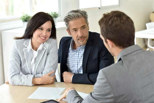 Une femme et un homme sont assis côte à côte à une table et écoutent attentivement un autre homme en face d’eux. Tous les trois sont habillés en tenue professionnelle. La table est recouverte de papiers suggérant une rencontre professionnelle ou une discussion sur le plafonnement des loyers.